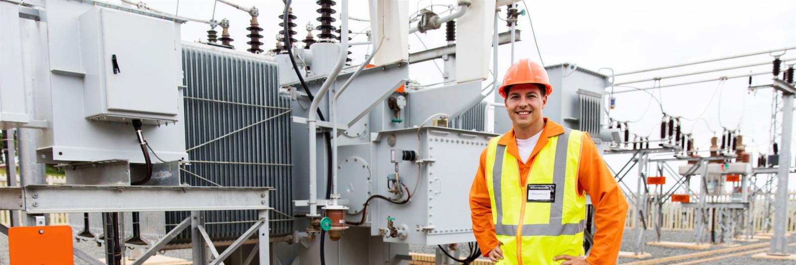 Elektrische Sicherheit & Energieversorgung garantiert Betreiber mit oranger Uniform & lachendem Gesicht steht vor einer elektrischen Kraftwerksanlage
